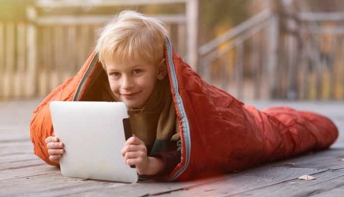 A small blond boy wrapped in a sleeping bag lies on the pier using a tablet.