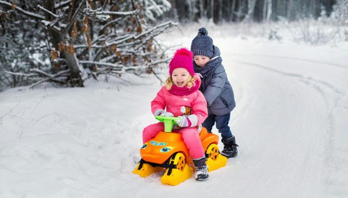 Boy in Blue Winter Clothes Pushing a Girl Riding on Toy Car on Snow Covered Ground