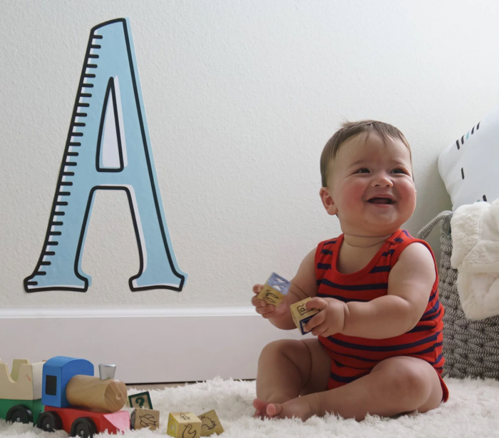 A baby sitting in front of a large letter A on his wall.